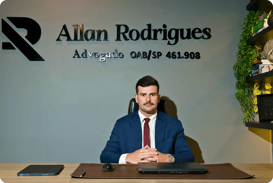 Allan Rodrigues - Advogado OAB/SP 461.908 em seu escritório, sentado à mesa com logotipo ao fundo